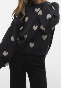 Pull en tricot bleu marine avec motifs de cœurs beiges, manches larges, col rond et coupe décontractée. Le matériau semble doux et texturé.