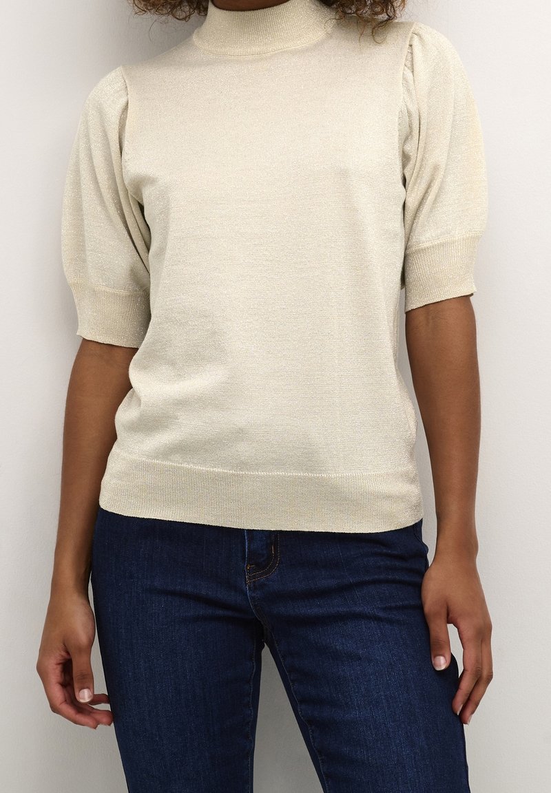 Femme portant un haut en maille crème à manches courtes bouffantes et un jean bleu foncé sur un fond uni.