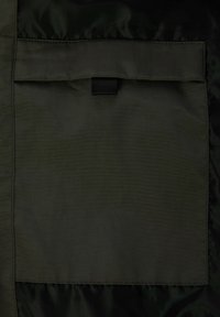 Tejido verde oscuro con una textura suave, presenta un bolsillo de parche alineado verticalmente en el centro, asegurado con una tira de nylon negra.