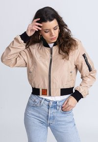 Licht beige bomberjack met een ritssluiting aan de voorkant, zwarte geribbelde manchetten en zakken met een flap, geëxposeerd tegen een neutrale achtergrond.