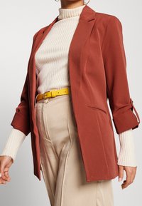 Rostfärgad kavaj i slät tyg, med skräddarsydd passform, slagkrage och uppvikta ärmar. Kombinerad med en ribbad crèmefärgad polo och ett gult bälte.