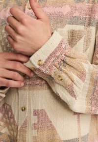 Mehrfarbige, strukturierte Web-Hemd mit geometrischen Mustern, hellbeige Grundfarbe und kontrastierenden rosa und braunen Akzenten. Die Knöpfe an den Manschetten sind sichtbar.