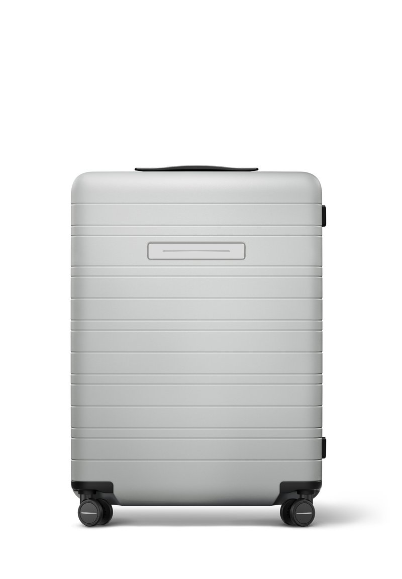 Valise rigide argentée avec des rainures horizontales, une poignée encastrée, quatre roulettes pivotantes noires et un emplacement pour une étiquette nom.