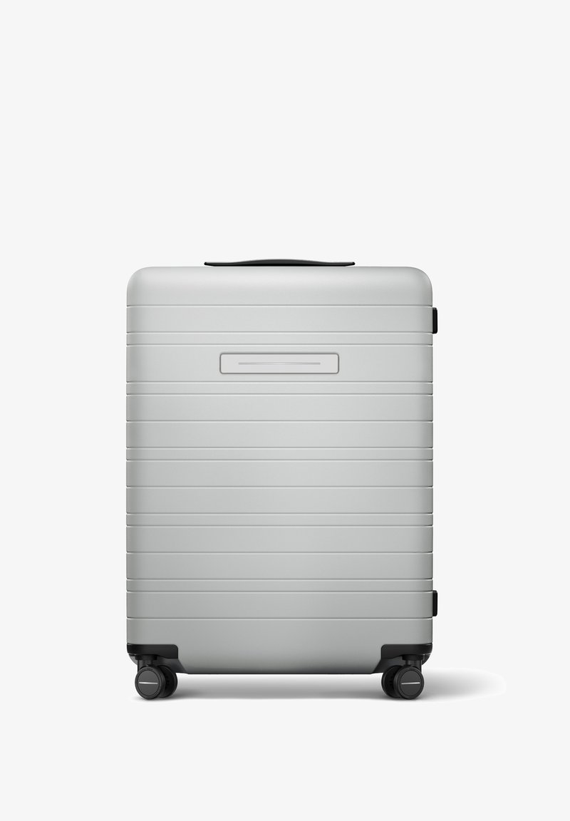 Valise rigide argentée avec des rainures horizontales, une poignée encastrée, quatre roulettes pivotantes noires et un emplacement pour une étiquette nom.