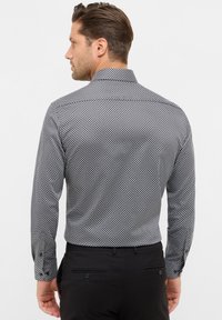 Homme portant une chemise à manches longues noire et blanche à motifs et un pantalon noir, se tenant de dos contre un fond uni.