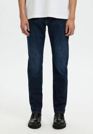 Personne portant un jean droit bleu foncé, des mocassins noirs et une chemise blanche sur un fond uni clair.