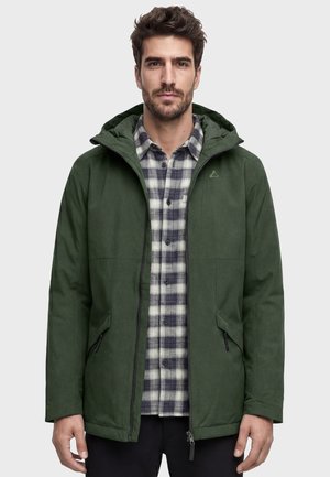 Mann trägt eine dunkelgrüne Kapuzenjacke über einem schwarz-weiß karierten Hemd mit Knöpfen, steht vor einem einfarbigen hellen Hintergrund.