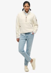 Pullover bianco in fetra con collo alto e zip a mezza lunghezza, dotato di tasche frontali. Abbinato a jeans azzurri chiari e sneakers beige.