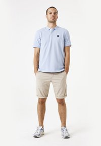 Lichtblauw poloshirt met een kraag en logo-patch, gecombineerd met beige opgerolde shorts. Model draagt grijze sportieve schoenen. Eenvoudige, casual outfit.