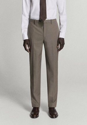 Hombre vistiendo pantalones claros marrones a medida, camisa blanca, corbata marrón oscuro y zapatos de cuero marrón oscuro, de pie frente a un fondo liso.