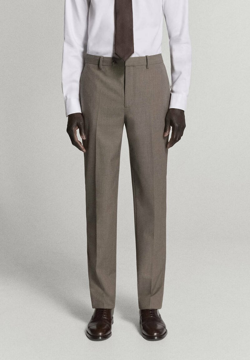 Homme portant un pantalon marron clair sur mesure, une chemise blanche, une cravate marron foncé et des chaussures en cuir marron foncé, debout devant un fond uni.