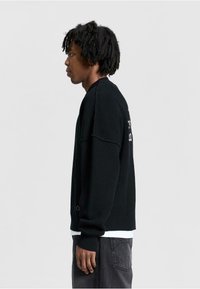 Black knitted sweater with a relaxed fit, ribbed texture, and oversized sleeves. Features embroidery on the back and layered with a white shirt.