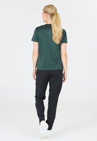 Mörkgrön kortärmad träningsskjorta tillsammans med svarta avsmalnande byxor och vita sneakers. Skjortan har ett mjukt, lätt tyg.