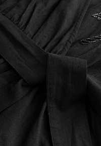 Tessuto nero con una finitura liscia, caratterizzato da un design arricciato e un prominente dettaglio cintura legata in vita. Minimalismo negli abbellimenti.
