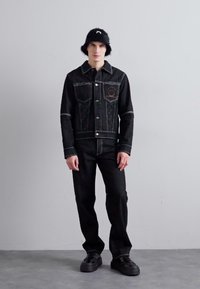 Musthave halli denim-jakk musta kontrastõmblustega ja kahe rinnataskuga, koos mustade pükstega ja karvase musta kübaraga.