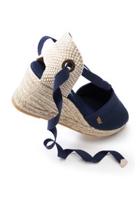 Scarpa con zeppa in tela blu navy con suola in juta intrecciata, cinturino alla caviglia in tessuto crema e lacci a nastro blu scuro. Dettaglio in metallo dorato sulla caviglia.