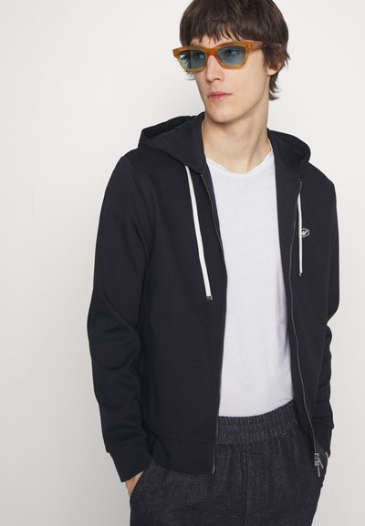 Black zip-up hoodie with white drawstrings, paired with a white t-shirt and textured dark pants. Blue-tinted rectangular sunglasses.