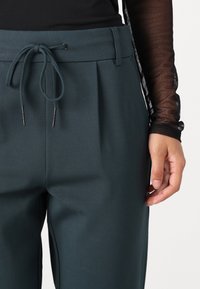 Tumedate roheline püksid volditud disainiga, elastse vöökoha ja nööriga. Sile kangas kerge läikega. Käsi toetub puusale.