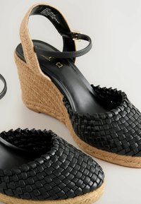 Sandali wedge in pelle intrecciata nera con tacco in juta naturale testurizzato e cinturino alla caviglia. Fodera interna liscia e dettagli hardware minimi.