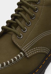 Κοντινή λήψη δερμάτινου μποτάκι σε ελιά πράσινο χρώμα, με πλεγμένα κορδόνια, μεταλλικά αυτιά κορδονιών, άσπρο ράψιμο και μαύρη σόλα με κίτρινες λεπτομέρειες ραφής.