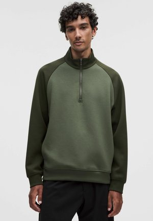 Man met krullend haar, gekleed in een olijfgroene trui met een kwartrits en zwarte broek, staand tegen een effen lichtgrijze achtergrond.