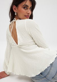 Witte, gestructureerde, lange mouwen top met een diepe V-rug en striksluiting. Geschulpte zoom. Gecombineerd met blauwe jeans.