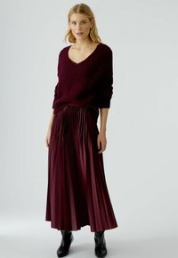 Burgunderfarbener Strickpullover mit V-Ausschnitt, kombiniert mit einem plissierten, bodenlangen Rock in passendem Burgunder. Schwarze Knöchelbots runden das Outfit ab.
