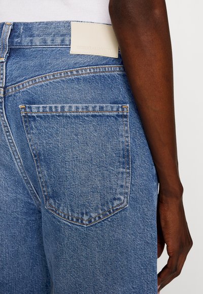 Lyseblå denimjeans har en baklomme med oransje søm og en rektangulær hvit lærpatch i midjen.