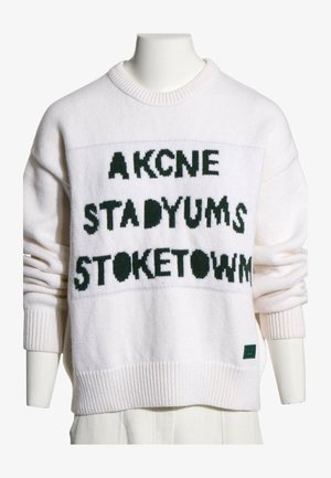 Pull tricoté blanc avec texte noir « AKCNE STADYUMS STOKETOWN » affiché à l'avant, porté sur un mannequin.
