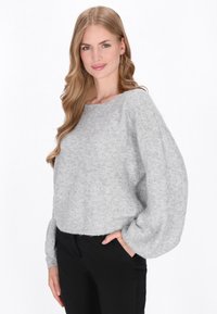 Kobieta z długimi blond włosami, nosząca kolczyki w kształcie kół, luźny szary sweter i czarne spodnie, stojąca z jedną ręką w kieszeni.