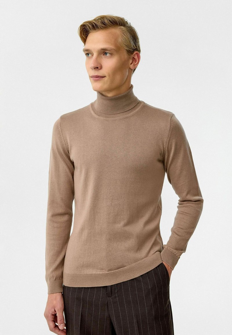 Coltrui in zacht beige breiwerk, aansluitend ontwerp met geribde halslijn en manchetten, gecombineerd met donker gestreepte broek.