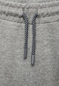 Šedé tepláky s texturovanou tkaninou, elastickým pasem a stahovací šňůrkou, doplněné tmavými tkanými detaily a plastovými špičkami.