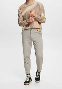 Beżowy sweter w abstrakcyjny wzór, jasnoszare spodnie oraz czarne trampki za kostkę z białymi sznurówkami. Dopasowany krój i gładka faktura materiału.