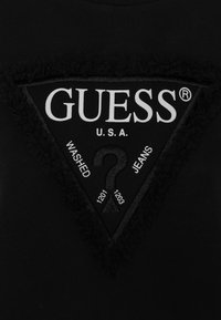 Černé tričko se štítkem v podobě černého trojúhelníku s bílým logem "GUESS", texturovanými okraji a tištěnými detaily o "OPRANÝCH DŽÍNECH".