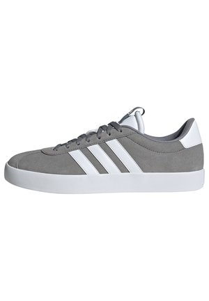 Zapatilla de ante gris con rayas blancas, punta redondeada, suela de goma y forro de tela. Presenta cordones gris y un lazo en el talón.