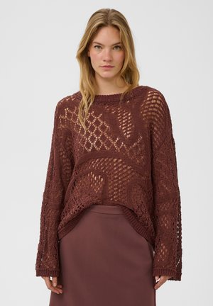 Femme aux longs cheveux blonds portant un pull ample marron en maille ajourée sur une jupe marron assortie, debout devant un fond uni.