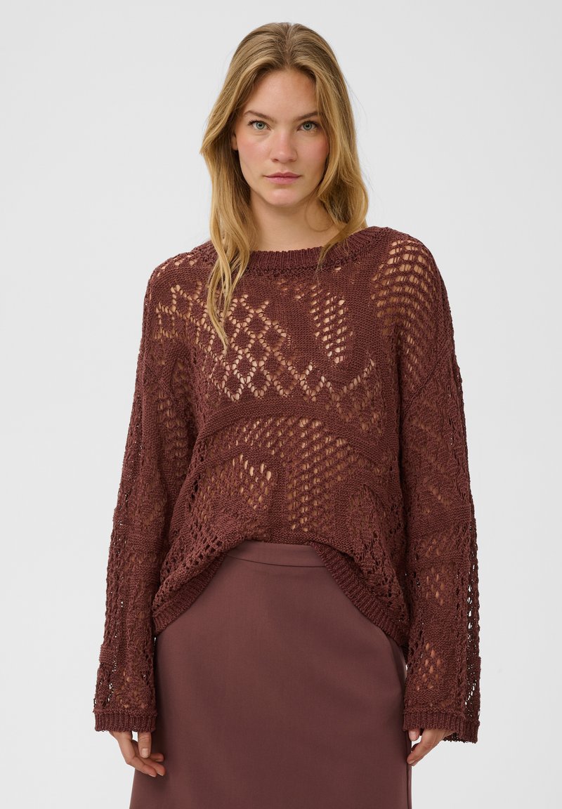 Femme aux longs cheveux blonds portant un pull ample marron en maille ajourée sur une jupe marron assortie, debout devant un fond uni.
