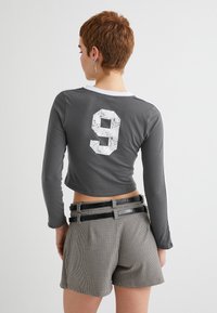 Grå langærmet cropped top med hvid nummer 9 trykt på ryggen. Bæres med mønstrede højtaljede shorts og kontrasterende sorte bælter.