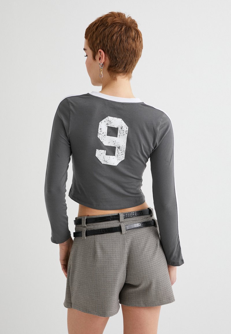Grå langærmet cropped top med hvid nummer 9 trykt på ryggen. Bæres med mønstrede højtaljede shorts og kontrasterende sorte bælter.
