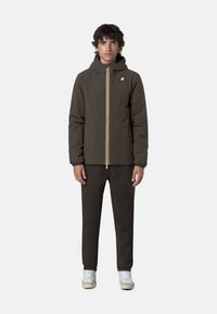 Giacca isolante verde scuro con cappuccio, dettagli della zip gialla e bianca, indossata sopra pantaloni marroni abbinati e sneakers bianche.