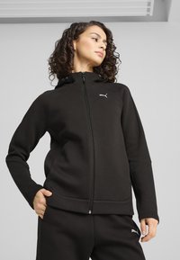 Chaqueta deportiva negra con cremallera y capucha, que presenta un logo de Puma, costuras estructuradas y una tela ligeramente texturizada. Combinada con pantalones a juego.