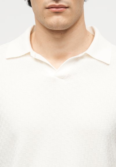 Νεαρός άνδρας που φοράει λευκό πουκάμισο πόλο με υφή και μαλακό γιακά, εμφανίζεται από τον λαιμό μέχρι το μέσο του στήθους σε ουδέτερο φόντο.