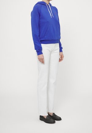 Osoba ubrana w niebieską bluzę z kapturem, białe spodnie o prostych nogawkach oraz czarne mokasyny, stojąca na tle jednolitego jasnego tła.