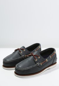 Scarpe da barca in pelle nera con cuciture bianche, lacci in pelle marrone e suola marrone chiaro. Presentano un design a scarpa bassa e punta rotonda.