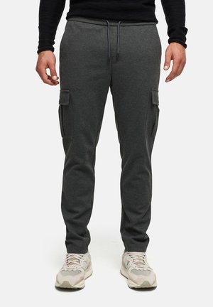 Uomo che indossa pantaloni cargo jogger grigio scuro e scarpe da ginnastica bianche, in piedi su sfondo bianco uniforme.