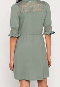 Femme portant une robe vert sauge avec des détails en dentelle sur le haut du dos et les manches, manches mi-longues bouffantes avec des poignets à volants, debout de dos.