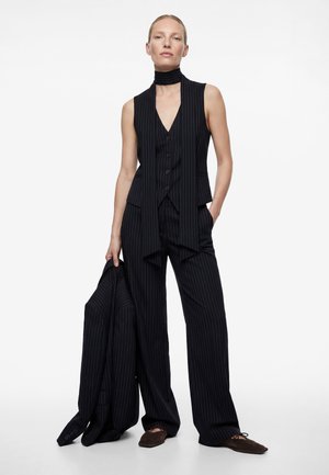 Femme portant un gilet sans manches à fines rayures noires et un pantalon large, tenant une veste assortie dans une main, debout devant un fond blanc.