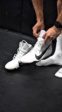 Scarpe sportive bianche con accenti neri, caratterizzate da un design alto, materiale testurizzato e lacci rotondi, posizionate su una superficie nera.
