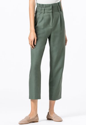 Pantalons taille haute verts avec plis, jambes fuselées et poches latérales ; en tissu lisse ; associés à des mocassins beiges.