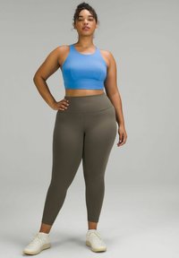 Blauwe sportbh en olijfgroene leggings, beide gemaakt van rekbaar materiaal. Model staat met een hand op de heup, met witte sportieve schoenen aan.